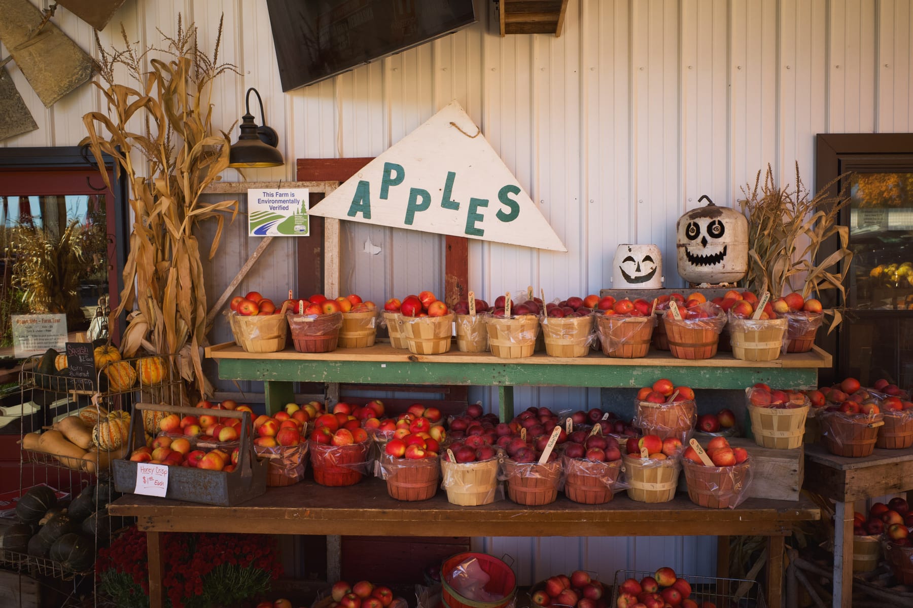 Baskets of red apples at a farm stand, with white tin jack-o-lanterns, underneath a sign with staggered letters saying APPLES