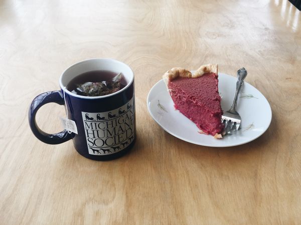 Tea bag steeping in navy blue Michigan Humane Society mug. Next to it: sweet beet pie with fork on a round white plate.