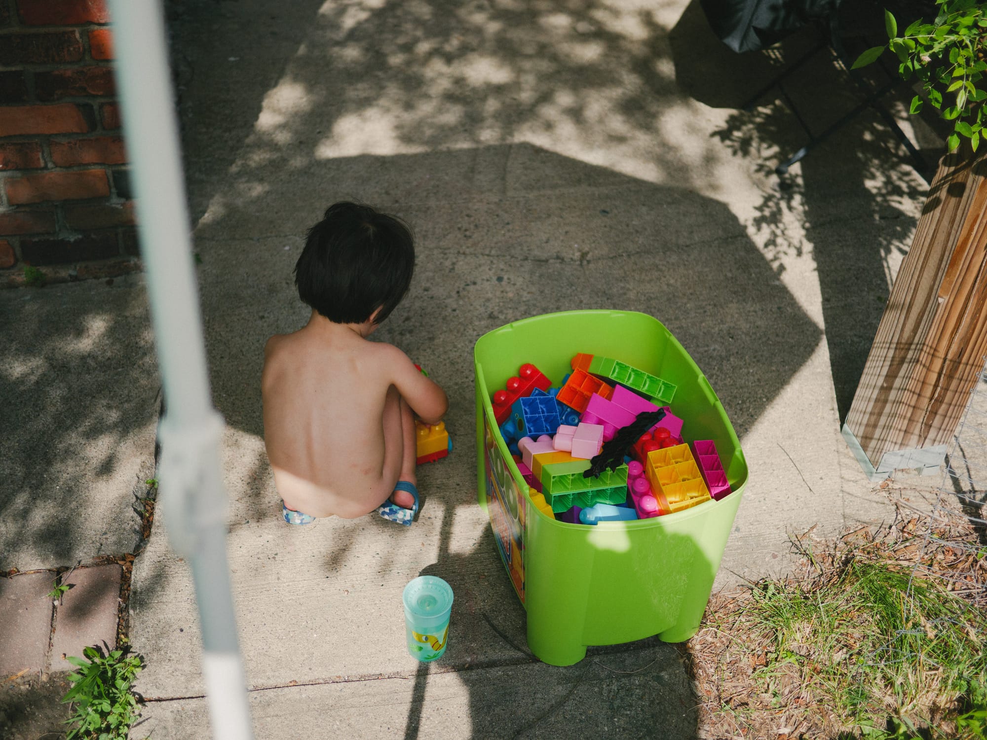 Rufus crouches shirtless on a sunlit concrete patio beside a bright green bin overflowing with colorful toy blocks.