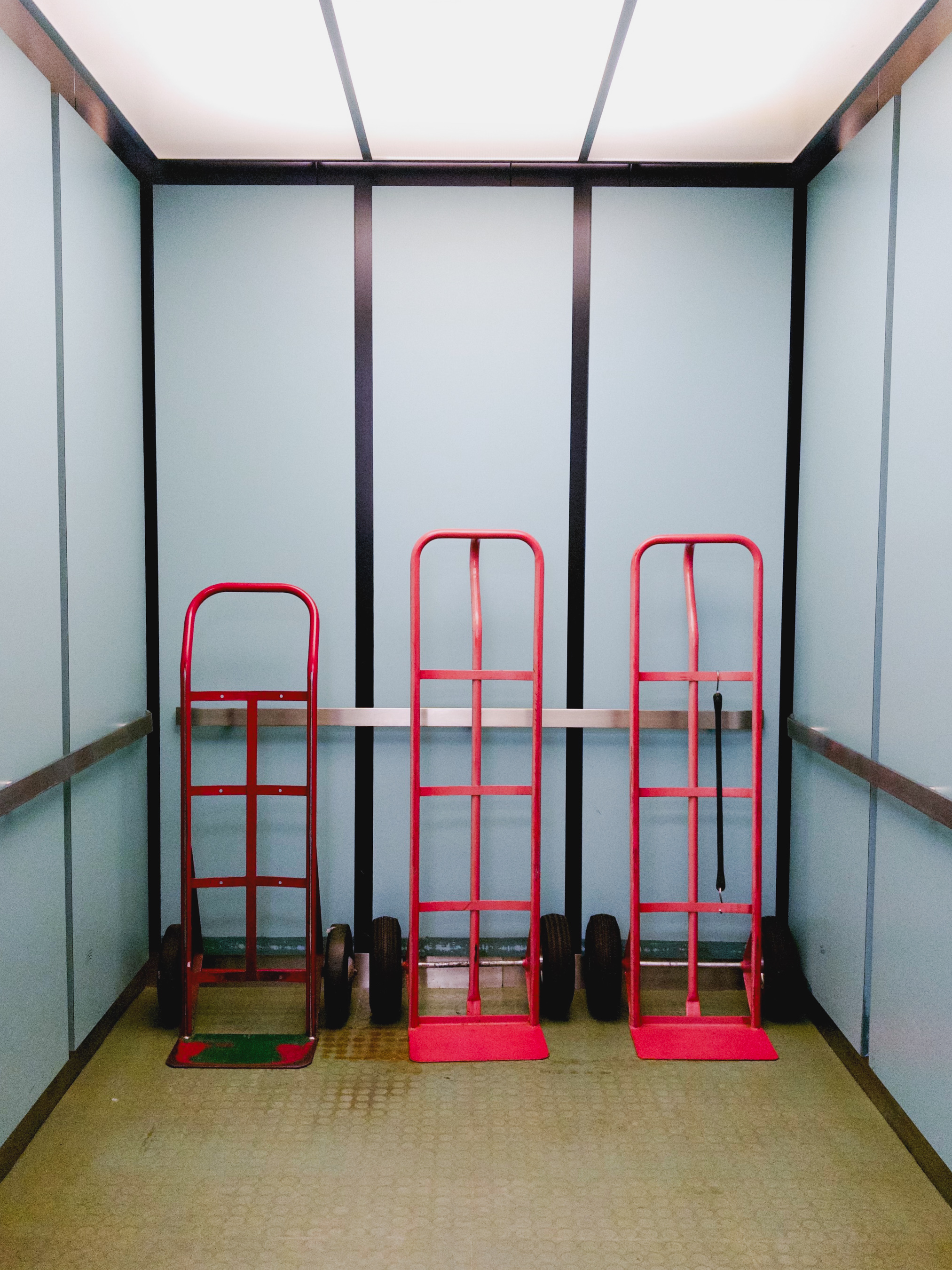 Three red hand trucks parked in a row inside a freight elevator with pale blue walls and a green floor.