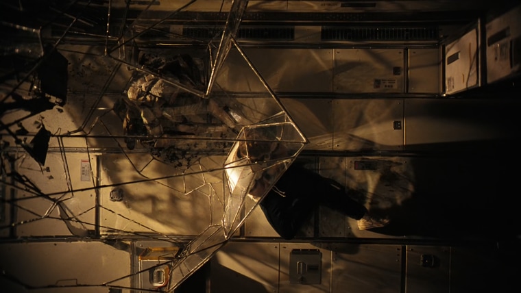 Person sitting on the floor of a spacecraft module beside a tall crystalline lattice of metal rods and translucent panels, lit by warm golden light with prismatic highlights on the structure and paneled walls.