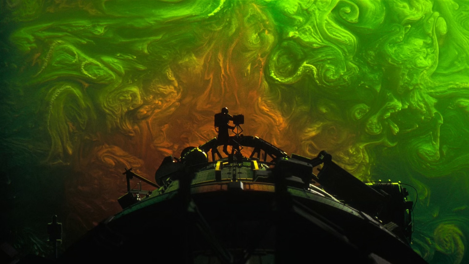 Silhouette of an astronaut standing on the hull of a spacecraft against a vast swirling nebula of orange and green cosmic clouds.