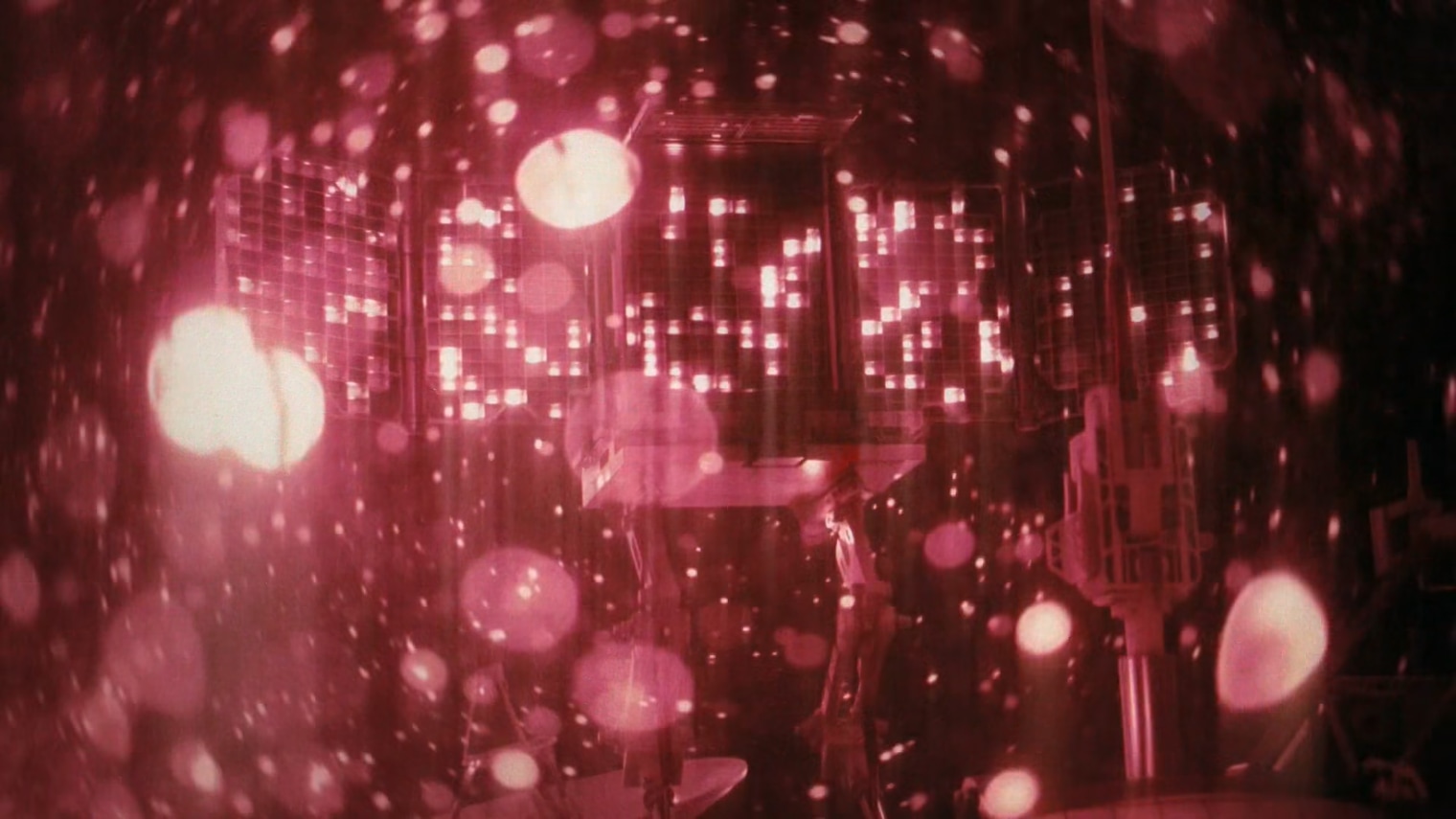 Close view of a spacecraft instrument panel with three gridded sections of small glowing lights, seen through heavy pink-red bokeh and soft lens flare.
