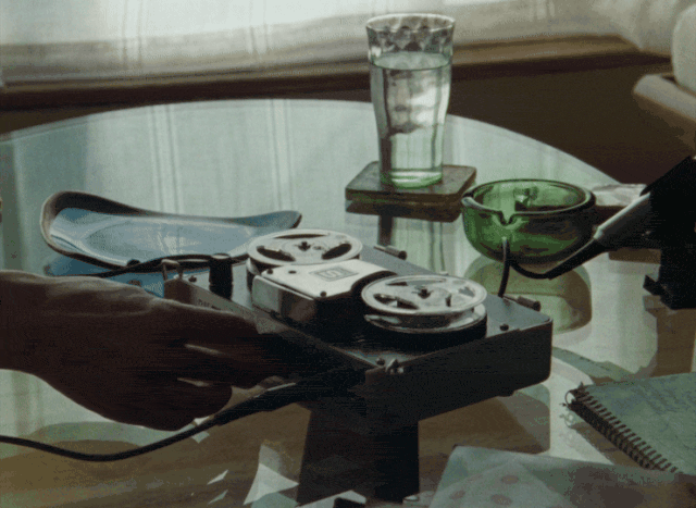 A hand adjusting a small portable reel-to-reel tape recorder on a glass coffee table, its reels turning, beside a glass of water, a green glass ashtray, and a pair of sunglasses.